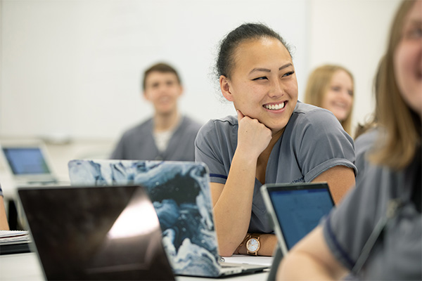 Smiling student in scrubs sits at a classrom desk with a computer