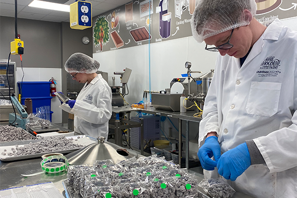 Two students in sterile gear package chocolate in an industrial kitchen.