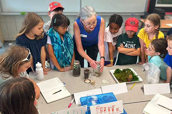 A group of children stand around a table while an instructor teaches using soil and plants in glass jars
