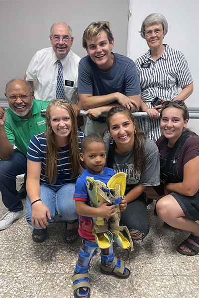 A young pediatric patient hold his old leg prosthetics while wearing his new ones with Shalese Evertsen, Alex Bushman, Kate Ruff, John Ruff, a clinic director, and two senior LDS missionaries.